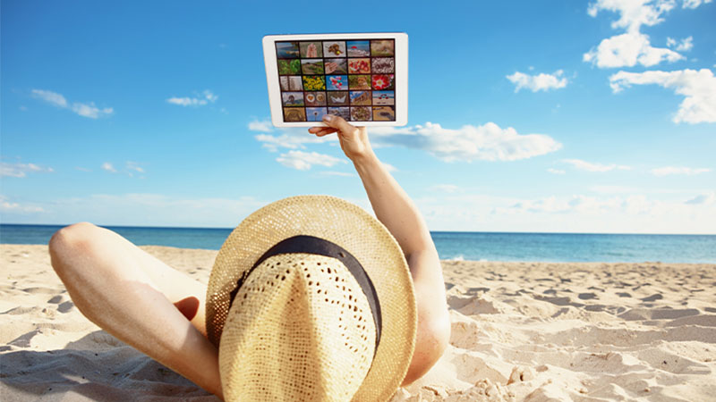 Pessoa deitada na areia da praia com chapéu de palha a ver TV no tablet