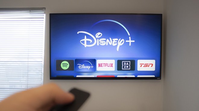 Mão a segurar num telecomando e a operar um televisor inteligente, montado na parede junto a uma janela, com diversas aplicações no ecrã, destacando-se a da Disney+