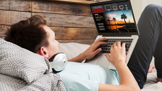 Rapaz deitado numa cama, com headphones no pescoço, a fazer streaming de vídeo num computador portátil pousado nos joelhos