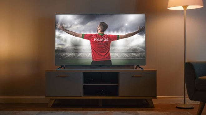 Televisão sobre uma mesa, numa sala com iluminação fornecida por um candeeiro de pé. No ecrã, pode ver-se um apoiante da seleção portuguesa de futebol, de costas e de braços abertos, virado para uma arena