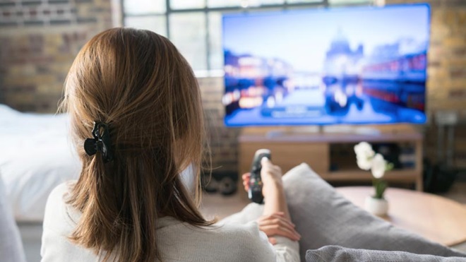Mulher sentada num sofá, vista de costas, com um telecomando na mão para usar um televisor