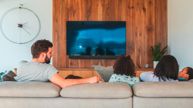 Família sentada no sofá da sala, de costas, a ver televisão