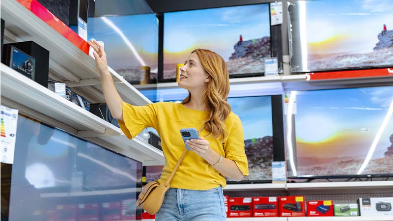 Pessoa numa loja de eletrónica observa televisores expostos numa parede, segura um smartphone e alcança uma prateleira com aparelhos.