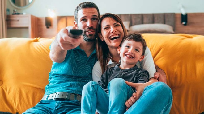 Pai, mãe e filho de cerca de 6 anos vêem televisão no quarto, em frente a uma cama de casal com colcha amarela. O pai segura na mão o comando
