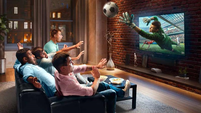 Grupo de amigos homens vê futebol numa televisão em casa; a imagem de jogador a segurar a bola sai do ecrã