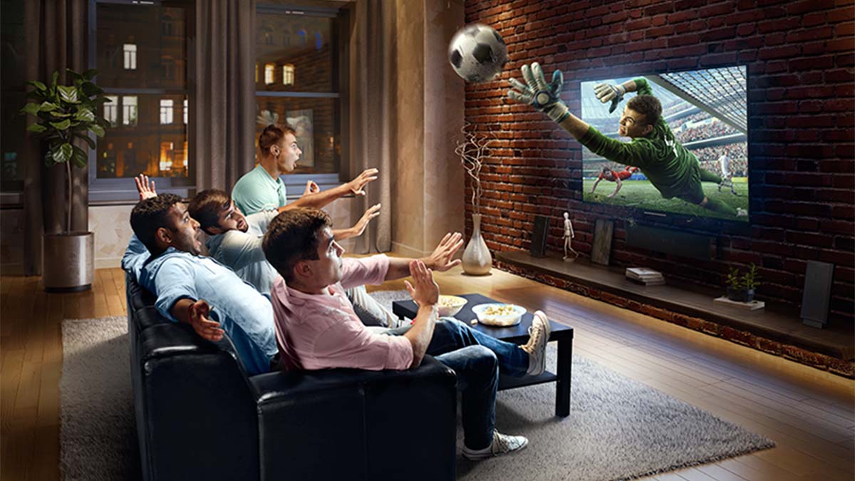 Grupo de amigos homens vê futebol numa televisão em casa; a imagem de jogador a segurar a bola sai do ecrã