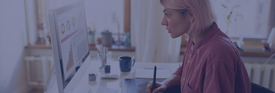 Mulher a olhar para o ecrã de um computador, onde se veem gráficos, enquanto toma notas num papel que se encontra em cima da secretária
