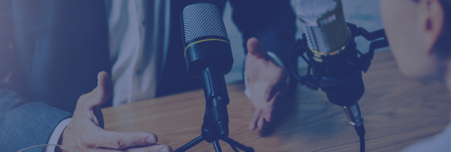 Duas pessoas sentadas a uma mesa com dois microfones pousados sobre a mesa. Ambas gesticulam